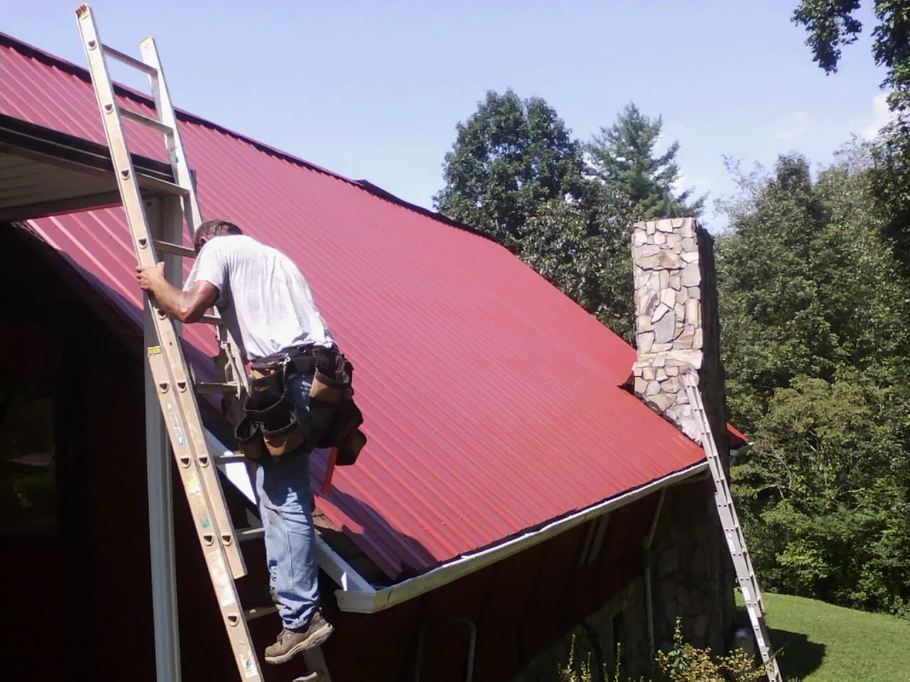 Roofer on a ladder beside a red metal roof and stone chimney — Soffit & Fascia Repair by Roofing Brothers in Perkiomenville
